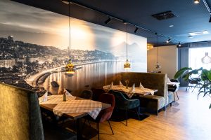 Sitzbereich im Restaurant mit großflächiger Fototapete einer nächtlichen Küstenstadt mit Berg. An den Tischen liegen braun-weiß karierte Decken. Es gibt grünliche Polsterbänke und dunkle Stühle.