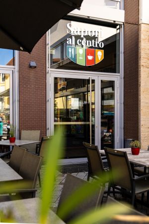 Verglaster Eingangsbereich des Restaurants "Sapori al centro" mit Logo über der Tür. Im unscharfen Vordergrund Tische und Stühle der Außengastronomie.