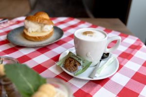 Tasse mit Milchschaumgetränk auf einer Untertasse mit Löffel, verpacktem Keks und Zuckerstick auf einer Rot-weiß karierten Tischdecke.