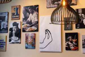 Wanddekoration bestehend aus mehreren gerahmten Bildern mit Italien-Bezug, darunter Motive von Spaghetti essenden Personen, einer italienischen Gasse und einer Skulptur, die Pizza isst.