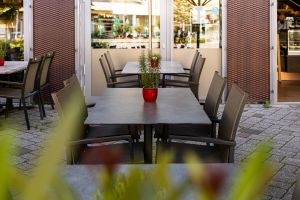 Ein grauer Terrassentisch mit einem roten Topf voller Rosmarin, umgeben von braunen Korbstühlen. Dahinter befindet sich eine geöffnete gläserne Eingangstür zu einem Restaurant.