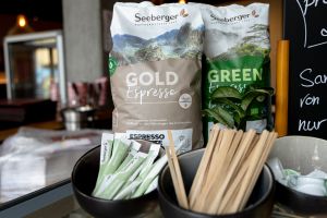 Zwei Kaffeebohnen-Packungen der Marke Seeberger hinter kleinen Schalen mit portionierten Zuckersticks, Holzrührstäbchen und Kaffeesahne-Kapseln.