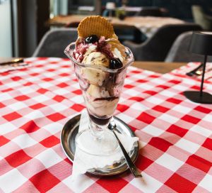 Ein hoher Eisbecher mit Vanilleeis, roter Fruchtsoße, Sahne, einer Kirsche und einer Eiswaffel auf einer rot-weiß karierten Tischdecke.
