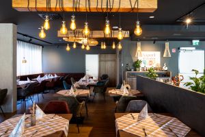 Restaurant-Innenbereich mit braun-weiß karierten Tischdecken und dunklen Polsterstühlen. Von der schwarzen Decke hängen Lampen aus Holzbalken. Im Hintergrund das Logo "Sapori al centro" an der Wand.