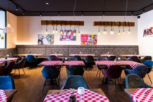 Innenraum eines Restaurants mit rot-weiß karierten Tischdecken auf quadratischen Tischen. Graue Polsterstühle und Sitzbänke. An der Wand hängen bunte Pop-Art-Bilder unter Holzbalken-Lampen.
