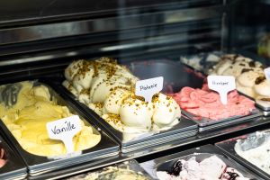 Eisvitrine mit Eissorten in Metallbehältern. Schilder zeigen Vanille, Pistazie mit gehackten Nüssen und Himbeer.