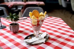 Eisbecher im Glas mit Waffel, Ananas- und Birnenscheiben sowie roter Sauce. Der Becher steht auf einem Metalltablett auf rot-weiß karierter Tischdecke.