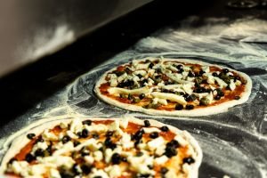 Zwei rohe Pizzen auf einer bemehlten dunklen Arbeitsfläche, belegt mit Tomatensauce, Mozzarellastreifen, schwarzen Oliven und Kapern.