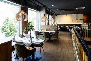 Heller Restaurant-Innenraum mit holzfarbenem Boden, Tischen mit karierten Decken und Polsterstühlen in Grün und Grau. Große Fensterfront, Holz-Hängelampen und ein hölzerner Raumteiler.
