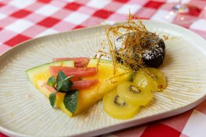 Ein Schokoladendessert mit einem filigranen Zuckergebilde, serviert mit Stücken von Melone, roter Grapefruit und Kiwi auf einem länglichen Teller.