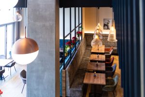 Lange graue Polsterbank mit Holztischen, getrennt durch eine halbhohe Wand mit Glasaufbau.