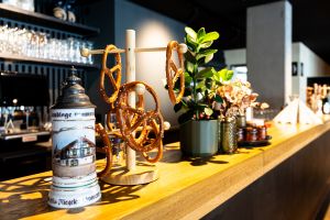 Hölzerne Theke, darauf ein traditioneller Bierkrug mit Zinndeckel und ein hölzerner Brezenständer mit aufgehängten Brezen. Daneben eine grüne Pflanze in einem Topf.