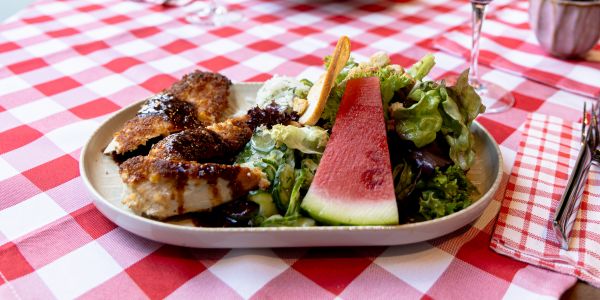 Teller mit panierten Fleischstreifen an dunkler Sauce und gemischtem Salat mit einer Scheibe Wassermelone.