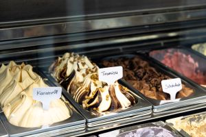 Eisvitrine mit verschiedenen Eissorten in Metallbehältern. Sichtbare Schilder beschriften die Sorten Karamell, Tiramisu und Schokolade.