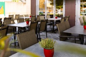 Nahaufnahme eines grauen Terrassentisches mit kleinem roten Pflanztopf mit Kräutern. Um den Tisch stehen braune Korbstühle. Im unscharfen Hintergrund weitere Tische der Außengastronomie.