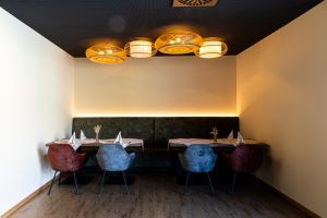Kleine Sitzecke in einem Restaurant mit grüner Polsterbank, zwei Tischen mit karierten Tischdecken und vier gepolsterten Stühlen. An der schwarzen Decke hängen vier runde Korblampen.