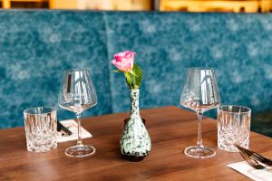 Eine rosa Rose in einer Vase zwischen zwei Weingläsern und Wassergläsern vor einer blauen Polsterlehne.