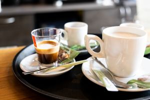Schwarzes Serviertablett mit einem Espresso im kleinen Glas und einem Heißgetränk mit Milchschaum in einer weißen Tasse. Auf den Untertassen liegen Löffel und verpackte Zuckersticks.