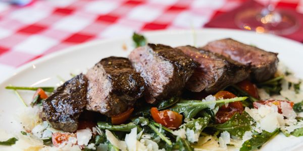 In dicke Scheiben geschnittenes, medium gebratenes Steak auf einem Bett aus Rucola, Kirschtomaten und gehobeltem Parmesan auf einem weißen Teller.