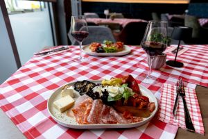 Vorspeisenteller mit Salami, verschiedenen Käsesorten, gegrillter Aubergine, Paprika und Bruschetta und daneben Rotwein, auf rot-weiß karierter Tischdecke.