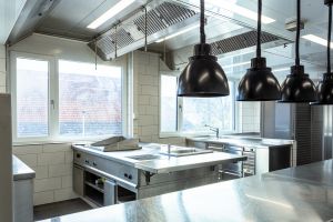 Gastro-Küche mit Edelstahlarbeitsflächen, Spülbereich, schwarzen Wärmelampen Fenstern.