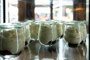 Mehrere kleine Gläser, gefüllt mit einem geschichteten Dessert aus dunklem Boden und einer hellen, weißen Creme, aufgereiht in einer gläsernen Vitrine.
