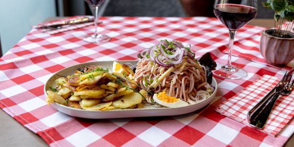 Teller mit Wurstsalat, roten Zwiebeln, Bratkartoffeln, Schnittlauch und gekochtem Ei und Gläser mit Rotwein auf einer rot-weiß karierten Tischdecke.