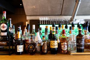 Theke mit zahlreichen aufgereihten Spirituosenflaschen, darunter Martini, Hennessy, Malibu und Chivas. Im Hintergrund ein grün beleuchtetes Regal.