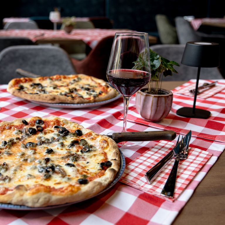 Zwei Pizzen mit schwarzen Oliven, daneben Gläser mit Rotwein, Besteck und eine kleine Pflanze im Topf auf einer rot-weiß karierten Tischdecke.