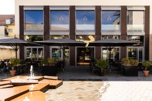 Vor dem Gebäude des Restaurants "Sapori al centro" mit großen Fenstern stehen Tische und Stühle unter schwarzen Sonnenschirmen. Im Vordergrund ist ein flacher Springbrunnen.