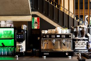 Kaffeestation unter einer Treppe mit Milchmaschine, Siebträgermaschine und Kaffeemühlen. Links ein grün beleuchtetes Regal mit Gläsern.