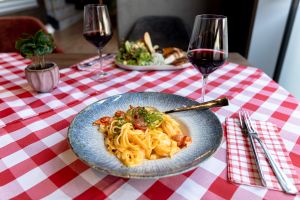 Pasta mit Kirschtomaten und Kräutern in einem tiefen Teller. Auf rot-weiß karierter Tischdecke, umgeben von Besteck, zwei Gläsern Rotwein und einer kleinen Pflanze. Im Hintergrund ein unscharfer Salat.