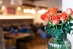 Rote Rosen in einer grünen Riffelglasvase vor einem warm beleuchteten im Gastraum.